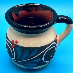 May include: A handcrafted ceramic mug with a cream-colored body, adorned with blue leaf and white swirl designs. The rim and handle are black, and the interior is a deep red. Red dots accent the design. The mug has a capacity of approximately 8 ounces.