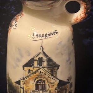 May include: Cream-colored ceramic pitcher with a handle and spout. The pitcher features a hand-painted illustration of a building and the word "EYGURANDE". The rim and handle are accented with orange and black details.