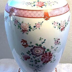 May include: A white ceramic vase with a wide body and a narrow neck. The vase is adorned with floral patterns in shades of pink, purple, and green. A pink band with a diamond pattern encircles the top.