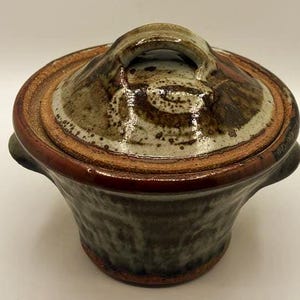 May include: A round, lidded ceramic pot featuring a mottled, earthy glaze in shades of brown, gray, and tan. The pot has two small side handles and a domed lid with a central arched handle, showcasing a textured, hand-thrown appearance throughout.