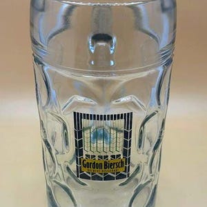 May include: A clear glass beer mug featuring a dimpled exterior surface and a sturdy handle. The front of the mug displays a rectangular logo with the text Gordon Biersch Brewery Restaurant in black and yellow lettering over a grid pattern.