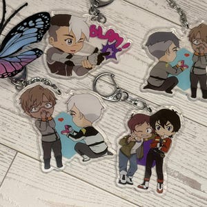 May include: Four clear acrylic keychains featuring anime-style character illustrations. One shows a character firing a weapon with the text "BLAM!". Two depict a character kneeling to propose with a ring. One shows two characters standing together.