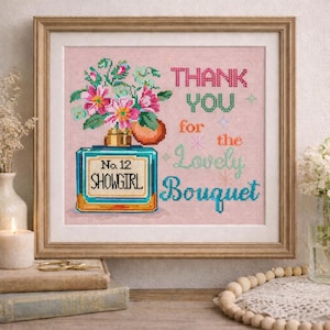 May include: Cross-stitch art in a wooden frame. The artwork features a perfume bottle with a floral bouquet and the text "THANK YOU for the Lovely Bouquet." The bottle is labeled "No. 12 SHOWGIRL."