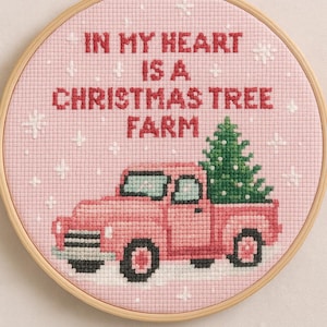 Può includere: Ricamo a punto croce in un telaio di legno. Il disegno presenta un camioncino rosa che trasporta un albero di Natale verde, con la scritta "IN MY HEART IS A CHRISTMAS TREE FARM" in rosso. Accenti di fiocchi di neve bianchi sono sparsi sullo sfondo rosa.