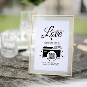 May include: A gold-framed sign with a white background and black text that reads "Share the Love".  The sign also features a black and white illustration of a camera with a QR code in the viewfinder.  The text below the camera reads "Scan the QR code to upload your photos! Help us relive the day through your lens."