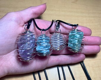 May include: Four crystal necklaces with different colored stones in silver wire cages. The necklaces are on black cords and are displayed on a hand.