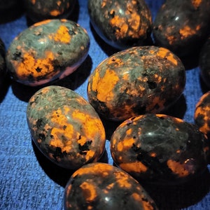 May include: A collection of oval-shaped stones with a dark, speckled appearance and vibrant orange patches. The stones are smooth and polished, with a natural, organic aesthetic. The background is a textured blue surface.