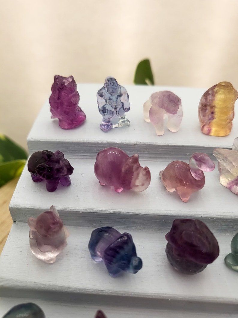 以下が含まれることがあります： カラフルな彫刻が施された小さな蛍石の動物の置物のコレクション。置物は白い段になったディスプレイスタンドに並べられています。置物には、ブタ、サイ、ヒツジ、クマ、鳥、猫が含まれています。