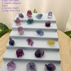 May include: A display of small, colorful crystal figurines, including a shark, penguin, frog, octopus, jellyfish, dolphin, and tortoise. The figurines are arranged on a tiered, light blue display stand. The crystals are various colors, including purple, blue, and yellow.