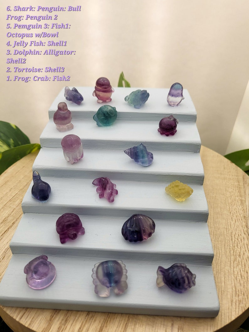 May include: A display of small, colorful crystal figurines, including a shark, penguin, frog, octopus, jellyfish, dolphin, and tortoise. The figurines are arranged on a tiered, light blue display stand. The crystals are various colors, including purple, blue, and yellow.