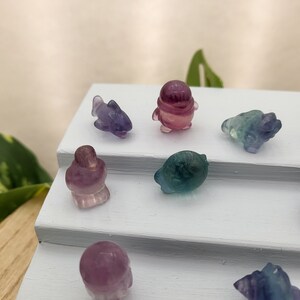 May include: A collection of small, carved gemstone figurines in various shapes, including a dolphin, penguin, and turtle. The figurines are primarily purple, green, and pink, and are displayed on a white, tiered surface. The overall theme is whimsical and decorative.