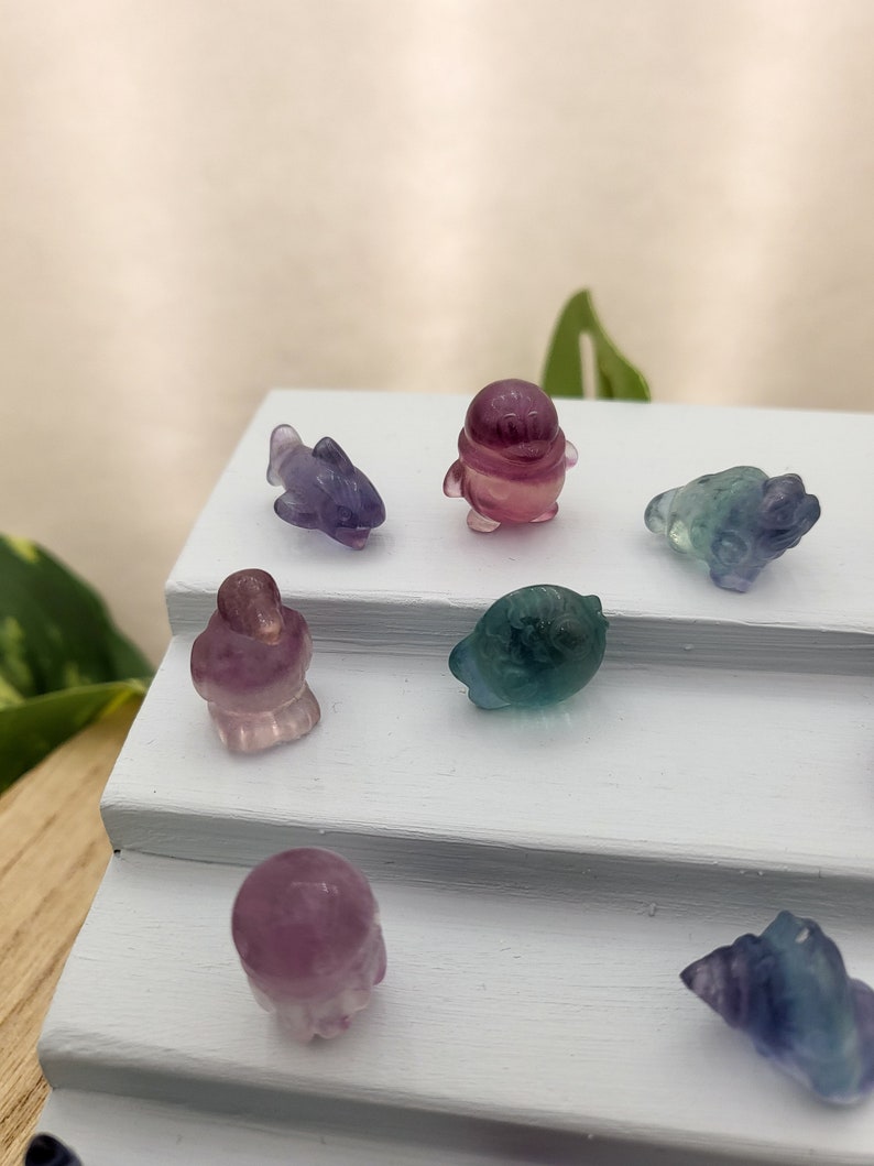 May include: A collection of small, carved gemstone figurines in various shapes, including a dolphin, penguin, and turtle. The figurines are primarily purple, green, and pink, and are displayed on a white, tiered surface. The overall theme is whimsical and decorative.