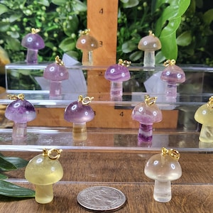 May include: A collection of 12 colorful crystal mushroom charms with gold accents. The charms are displayed on a tiered acrylic stand. The charms are a mix of purple, green, and yellow.