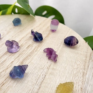 May include: A collection of small, carved gemstone figurines in various shapes, including an elephant, a seashell, and a dinosaur. The figurines are primarily purple, blue, and yellow, and are displayed on a light wooden surface.