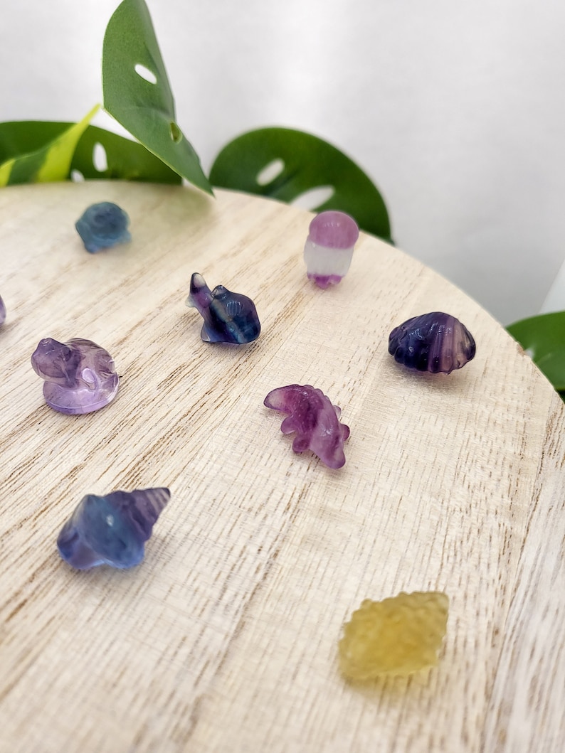 May include: A collection of small, carved gemstone figurines in various shapes, including an elephant, a seashell, and a dinosaur. The figurines are primarily purple, blue, and yellow, and are displayed on a light wooden surface.