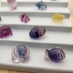 May include: A collection of small, carved gemstone figurines in various shapes, including a fish, crab, and seashell. The figurines are displayed on a tiered, light blue shelf. The gemstones have a translucent quality with purple, blue, and yellow hues.