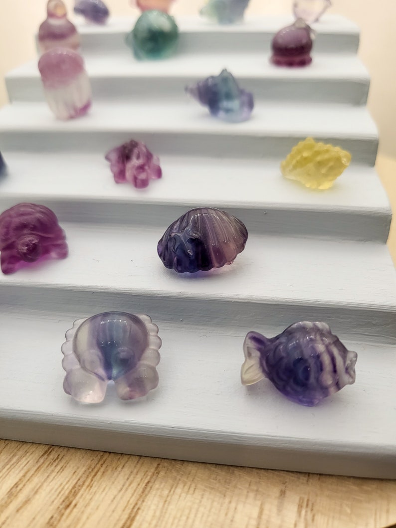 May include: A collection of small, carved gemstone figurines in various shapes, including a fish, crab, and seashell. The figurines are displayed on a tiered, light blue shelf. The gemstones have a translucent quality with purple, blue, and yellow hues.