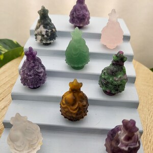 May include: A collection of nine small, carved crystal Christmas trees in various colors, including purple, green, pink, white, brown, and gray. The trees are arranged on a tiered display stand.