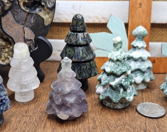 May include: A collection of seven small, carved, crystal Christmas tree ornaments. The trees are made of clear quartz, amethyst, green moss agate, and black and green jasper. The ornaments are arranged on a wooden surface.