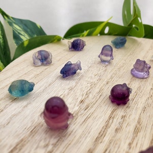 May include: A collection of small, colorful, carved gemstone figurines in various sea creature shapes, including fish, octopuses, and penguins. The figurines are shades of purple, blue, and green, displayed on a light wooden surface.