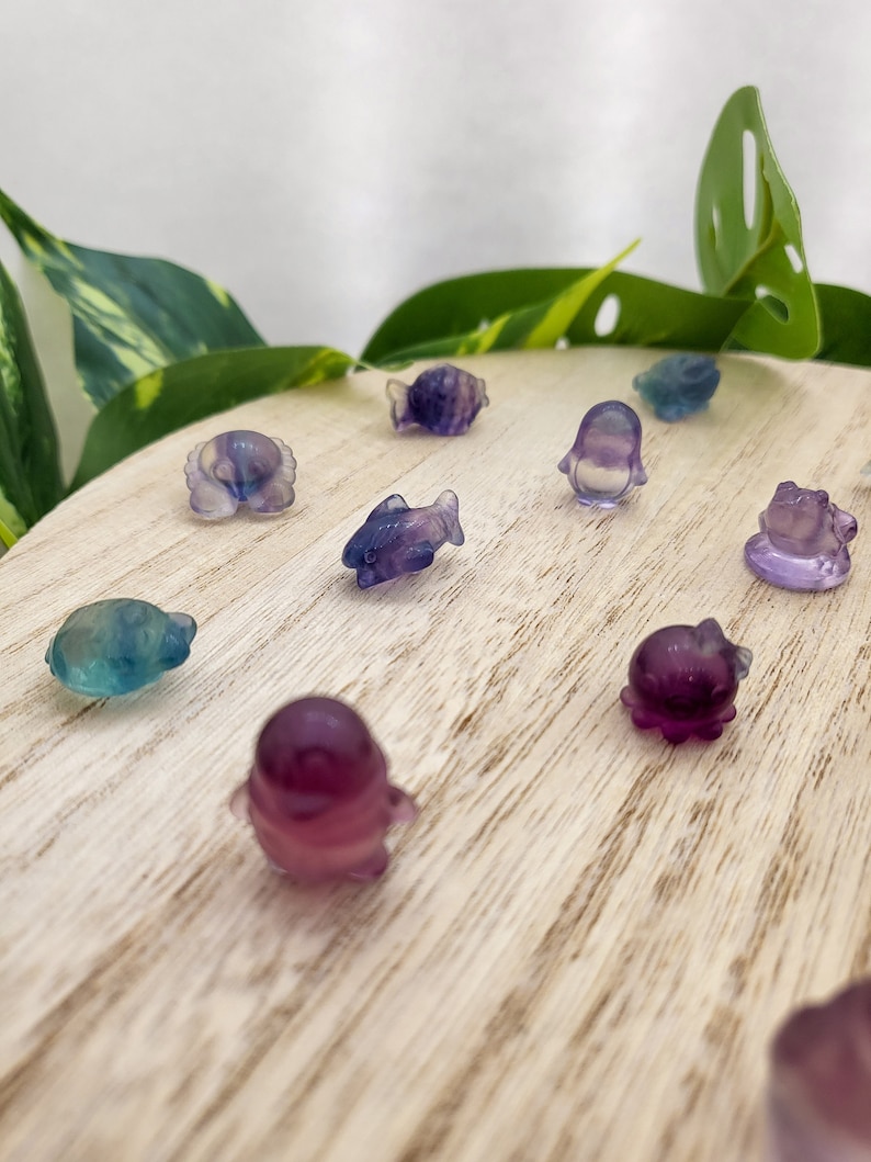 May include: A collection of small, colorful, carved gemstone figurines in various sea creature shapes, including fish, octopuses, and penguins. The figurines are shades of purple, blue, and green, displayed on a light wooden surface.