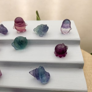 May include: A collection of small, colorful crystal figurines in various shapes, including animals and shells. The figurines are displayed on a white, tiered surface. The crystals feature shades of purple, blue, green, and yellow.