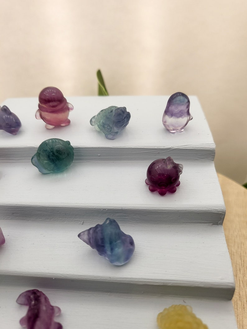 May include: A collection of small, colorful crystal figurines in various shapes, including animals and shells. The figurines are displayed on a white, tiered surface. The crystals feature shades of purple, blue, green, and yellow.