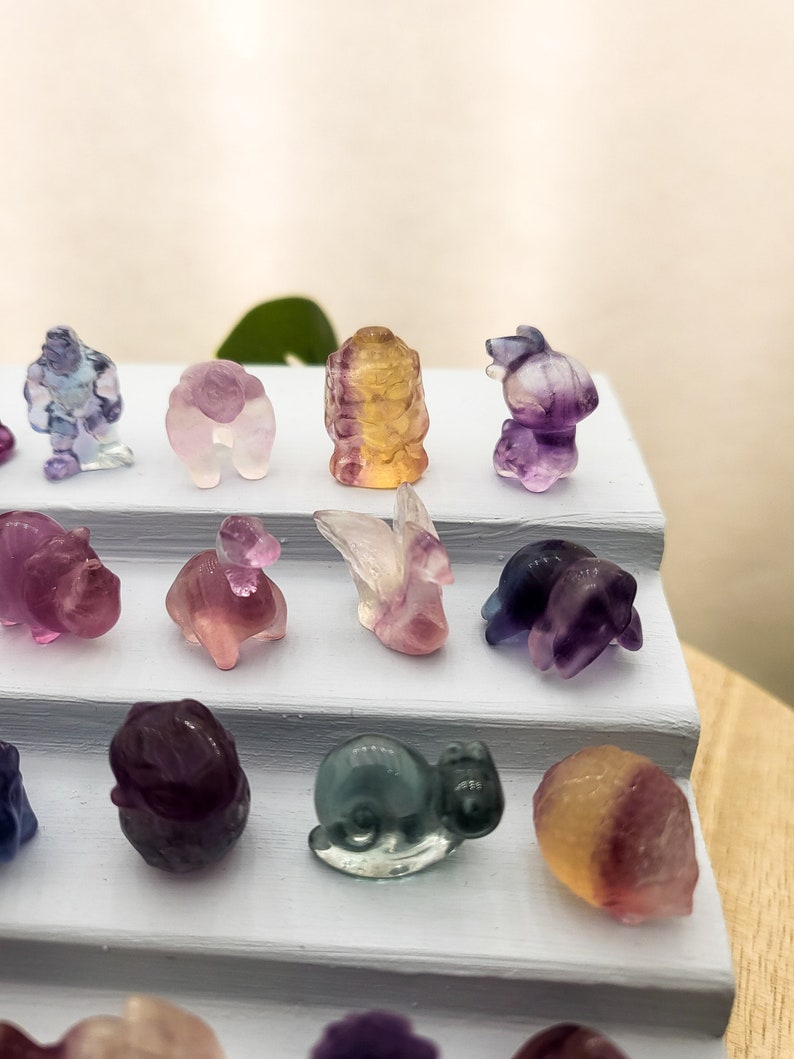 以下が含まれることがあります： 白い段になったスタンドに、小さくカラフルな彫刻された蛍石の動物の置物が並んでいます。置物には、白鳥、亀、カタツムリ、ウサギ、クマ、ドラゴンがあります。