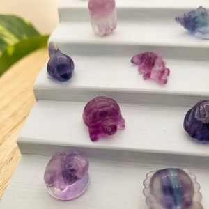 May include: A collection of small, carved gemstone figurines in various shapes, including animals and abstract forms. The figurines are primarily purple, with hints of blue and green, and are displayed on a white tiered stand.