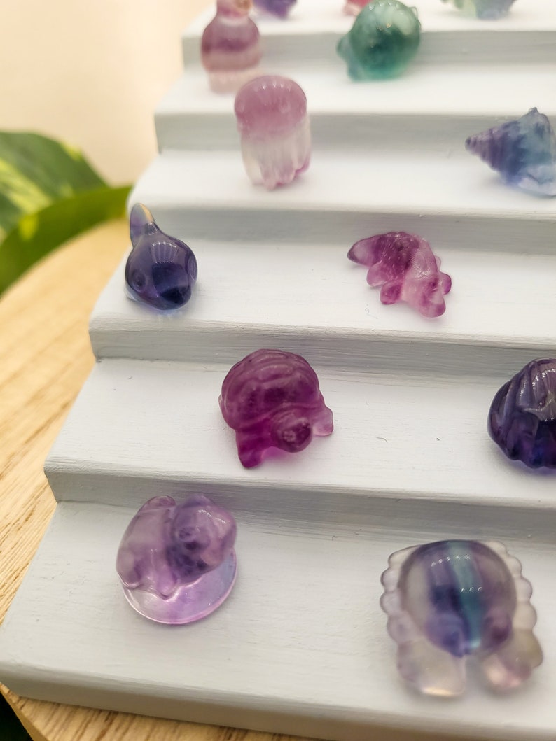 May include: A collection of small, carved gemstone figurines in various shapes, including animals and abstract forms. The figurines are primarily purple, with hints of blue and green, and are displayed on a white tiered stand.