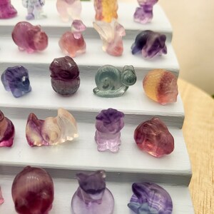 以下が含まれることがあります： 白い段になったスタンドに、小さくカラフルな彫刻された蛍石の動物の置物が並んでいます。置物には、カタツムリ、鳥、ブタ、ウサギ、猫、犬、カメ、魚、蝶、花があります。