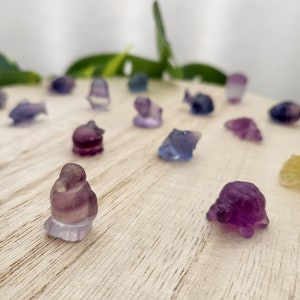 May include: A collection of small, carved gemstone figurines in various shapes, including fish, birds, and turtles. The figurines are primarily purple and blue, with some translucent areas. They are arranged on a light-colored wooden surface.