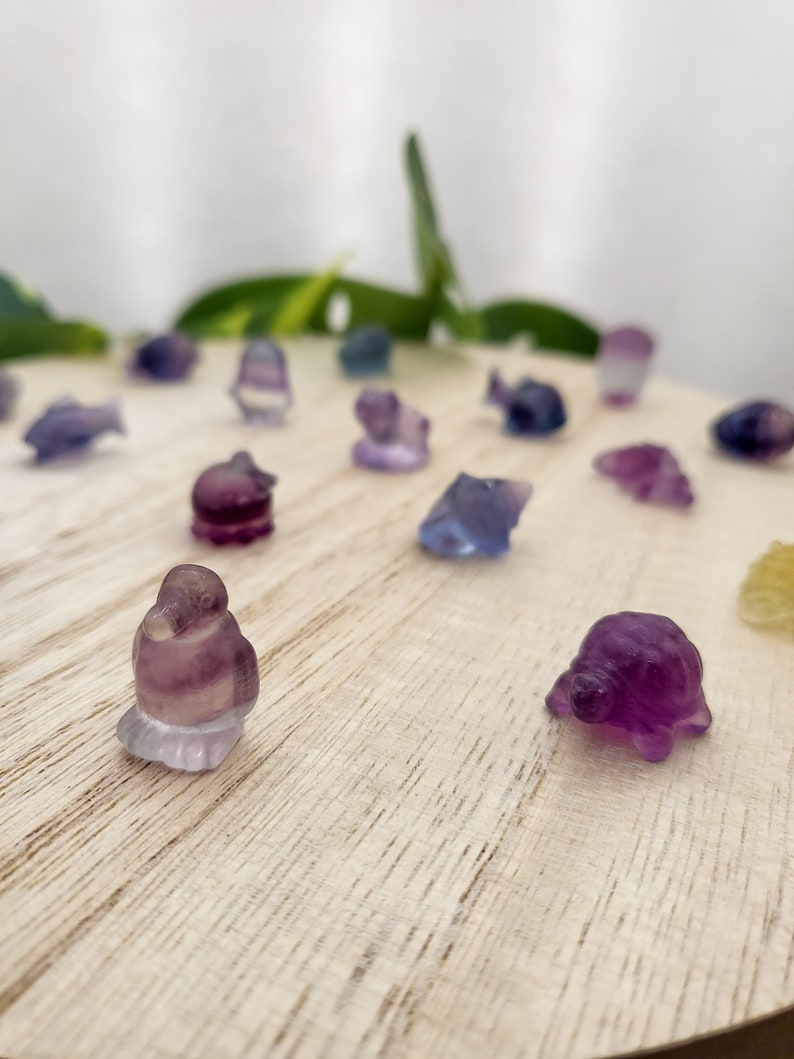 May include: A collection of small, carved gemstone figurines in various shapes, including fish, birds, and turtles. The figurines are primarily purple and blue, with some translucent areas. They are arranged on a light-colored wooden surface.