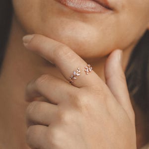 Puede incluir: Un anillo de oro rosa con un diseño de hojas, adornado con pequeñas piedras preciosas transparentes. El anillo envuelve el dedo, con los detalles de las hojas a ambos lados. La joya se muestra en la mano de una persona.