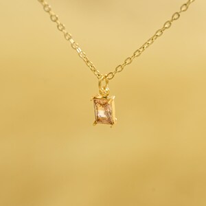 May include: A gold-colored necklace with a rectangular, light-brown gemstone pendant. The pendant is suspended from a delicate chain. The background is a solid, warm yellow color.