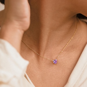 May include: A delicate gold necklace featuring a small, heart-shaped purple gemstone pendant. The necklace is worn on a person's neck, against a cream-colored shirt. The chain is fine and the pendant is centered.
