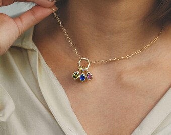 May include: A gold-colored necklace with a pendant featuring three colorful gemstones: green, blue, and pink. The necklace is being held up by a person wearing a light-colored shirt.