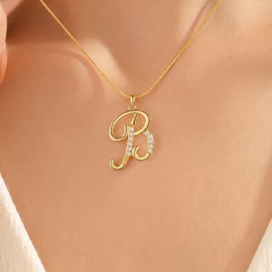 May include: A gold-colored pendant necklace featuring the letter "B" in a cursive font. The letter is embellished with small, sparkling stones. The necklace is displayed on a person's neck, showcasing the jewelry's design and style.