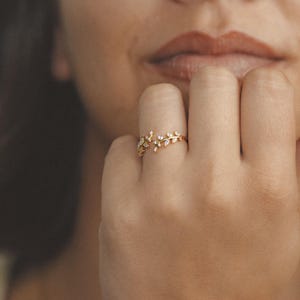 Op de afbeelding: Een goudkleurige ring met een bloemmotief, bezet met kleine, heldere edelstenen. De ring wordt aan een vinger gedragen, met de hand en een deel van het gezicht van een persoon zichtbaar. Het ontwerp van de ring lijkt op bladeren en kleine bloemen.