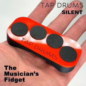 May include: A red and black rectangular musician's fidget toy with three black circular pads. The text "TAP DRUMS SILENT" and "The Musician's Fidget" are printed on the toy. The toy is held in a hand.