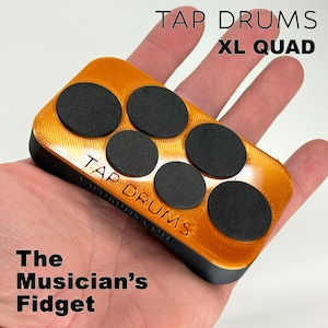 May include: Orange and black XL Quad Tap Drums fidget toy. The rectangular device has six black circular pads on an orange surface. The text "TAP DRUMS XL QUAD" is printed on the device. The text "The Musician's Fidget" is also visible.