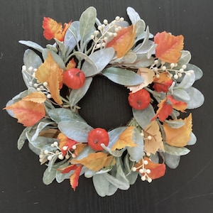 May include: A circular autumn wreath featuring sage green leaves, orange and yellow fall foliage, and small red pumpkins. The wreath is a decorative piece for the fall season.