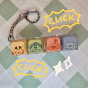 May include: A keychain featuring four square keycaps arranged in a row. Each keycap displays a different cartoon creature illustration in yellow, green, orange, and light blue. The word CLICK is written in yellow text twice with star accents.