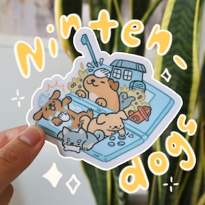 Könnte beinhalten: Ein Aufkleber mit einer Cartoon-Illustration von vier Hunden, die auf einem Nintendo DS spielen. Die Hunde sind ein brauner und weißer Hund, ein grauer und weißer Hund, ein brauner Hund und ein weißer Hund. Der Aufkleber ist auf weißem Hintergrund mit den Worten "Ninten-dogs" in Gelb.