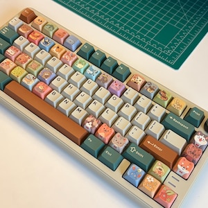May include: A mechanical keyboard featuring a light beige frame and a variety of colourful keycaps. The keys display a mix of standard letters, numbers, and symbols alongside various cartoon animal illustrations in shades of brown, green, and blue.