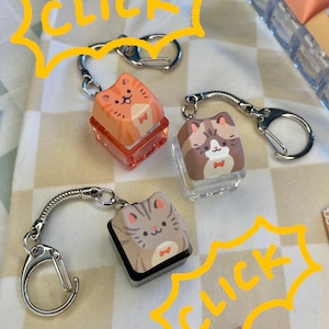 May include: Keychains with cat-themed keycaps. The keycaps are square and feature cartoon cat designs. The keychains have a silver metal chain and a clasp. The word "CLICK" is written in yellow in a speech bubble.