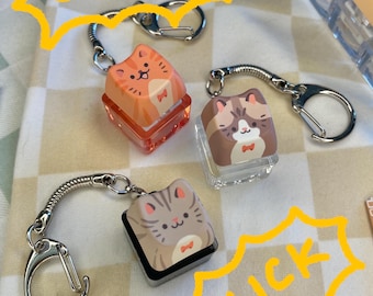 May include: Keychains featuring cat-themed keycaps. The keycaps are square and have a cartoon cat design. The keychains have a silver-colored metal chain and a clasp. The word "CLICK" is written in yellow in a speech bubble.