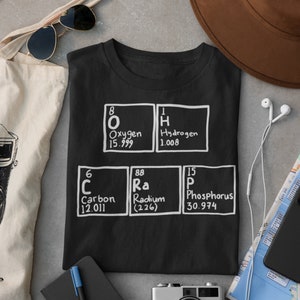May include: Black t-shirt with white text and images of chemical elements. The elements shown are Oxygen, Hydrogen, Carbon, Radium, and Phosphorus. The atomic number and weight are listed below each element.