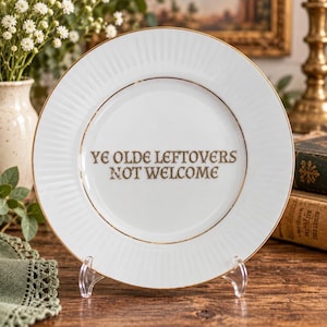 May include: A white ceramic plate with a gold-trimmed rim and a ribbed edge design. The centre of the plate features the text "YE OLDE LEFTOVERS NOT WELCOME" printed in a dark, stylised font. The plate is displayed upright on a clear stand.