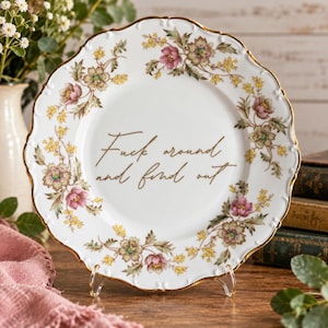 Puede incluir: Un plato de cerámica blanca con borde festoneado y acabado dorado, que presenta un patrón floral de flores rosas y amarillas alrededor del borde. El centro del plato muestra el texto "Fuck around and find out" en letra cursiva marrón.